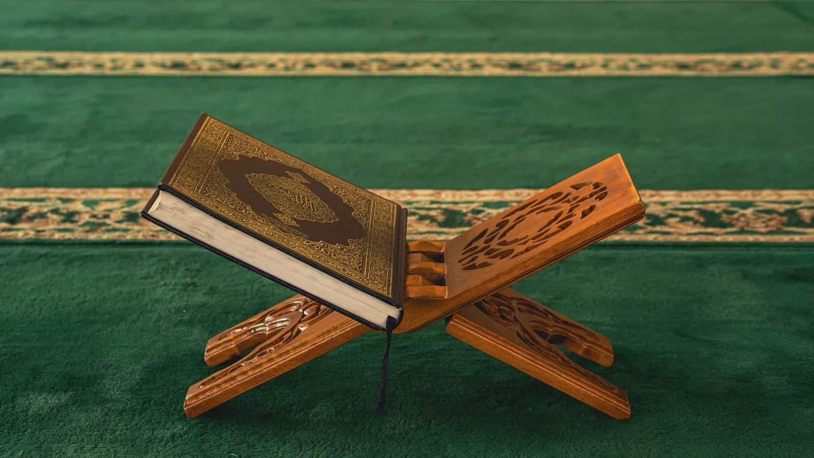 Un Coran fermé, à la couverture dorée, posé sur un porte-Coran en bois sculpté, sur un tapis de mosquée vert