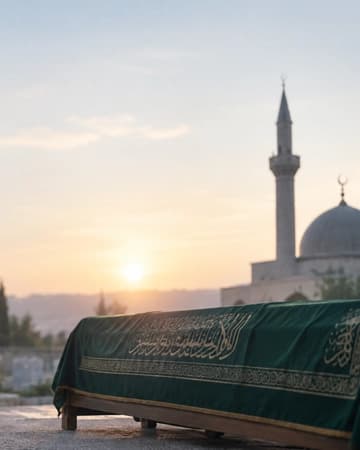 Un cercueil recouvert d’un drap vert calligraphié devant une mosquée, dans la lumière du soleil bas